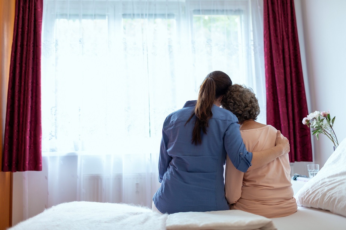 Female Home Nurse Hugging Elderly Woman on Bed. Back Wiev of Female Nurse With Her Arm Around Elderly Patient Shoulder.